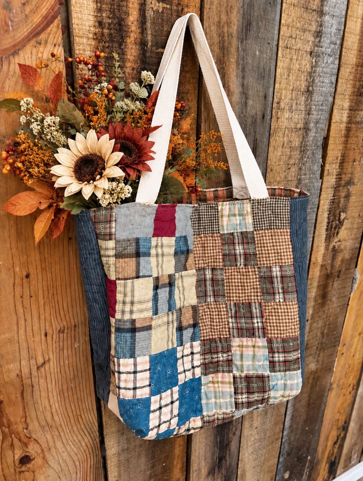 Quilted patchwork tote bag of blue, burgundy, and earth-toned squares with indigo side panels and a rust lining, holding an autumn flower arrangement