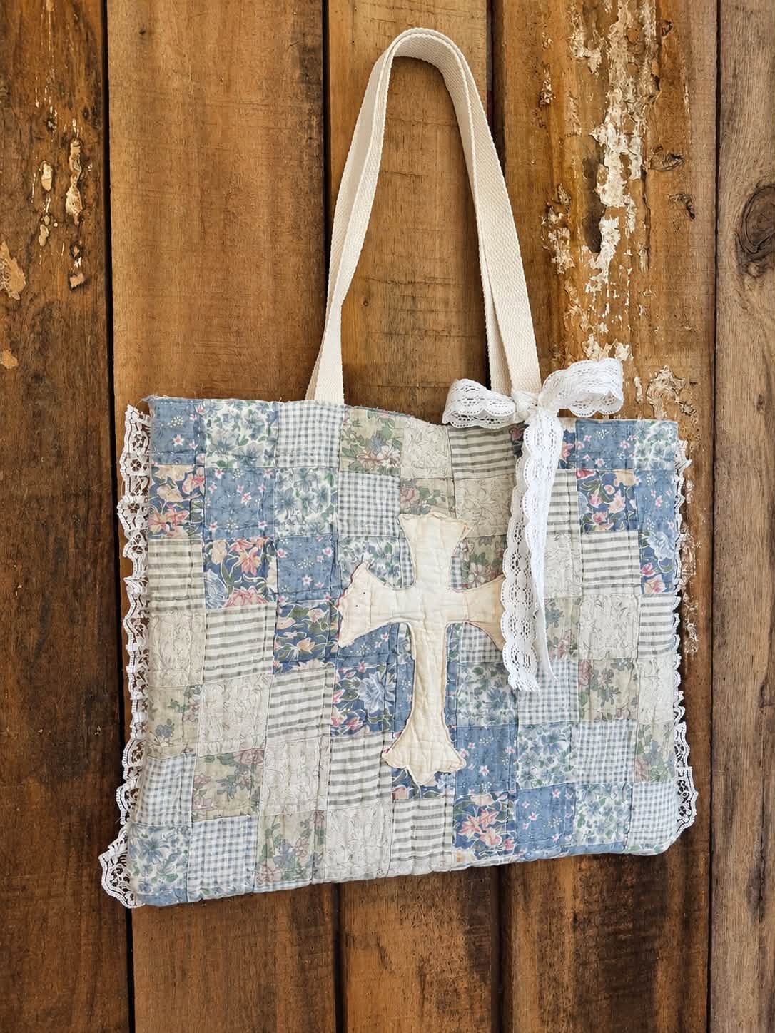 Patchwork tote bag in dusty blue, cream, and sage squares with lace edging, a stitched cross appliqué at the center, and a lace bow at the handle